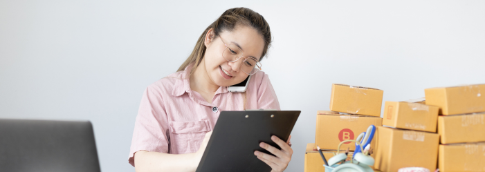 Woman smiling with a clipboard, a laptop, and stack of boxes to show an employee being productive.